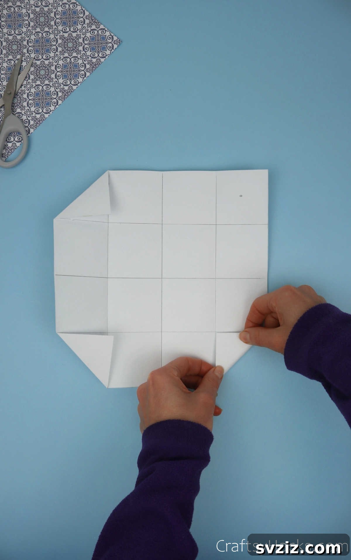 One corner of the grid-patterned paper being carefully folded inwards to form a small triangular flap, precisely aligned with the existing grid lines.