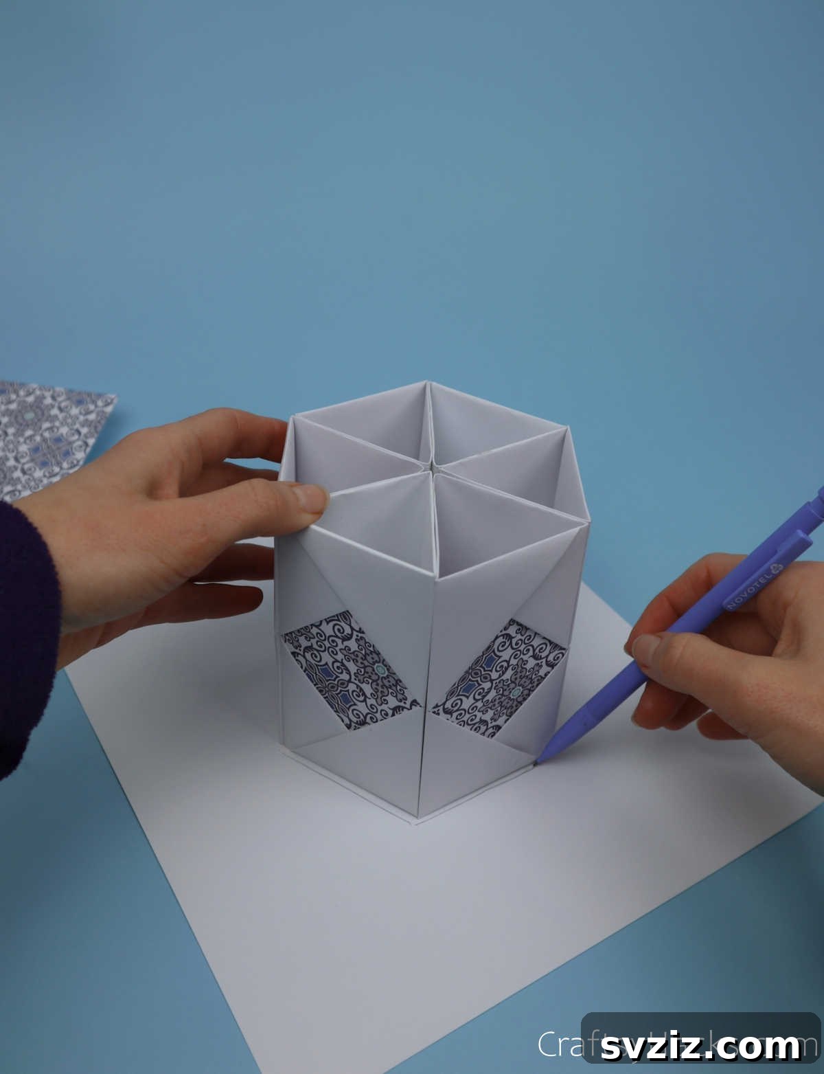 The assembled hexagonal pencil holder body placed on a sheet of thick paper, with a pencil tracing its exact outline to create the base template.