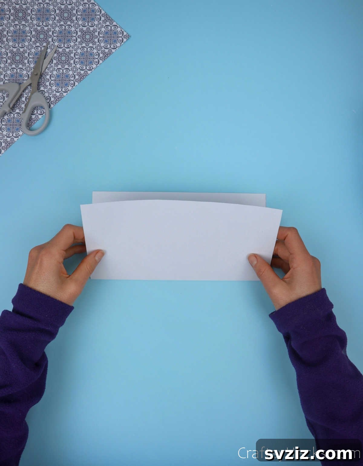 A square sheet of paper being folded horizontally in half, with attention to aligning the edges, to create a sharp central crease.