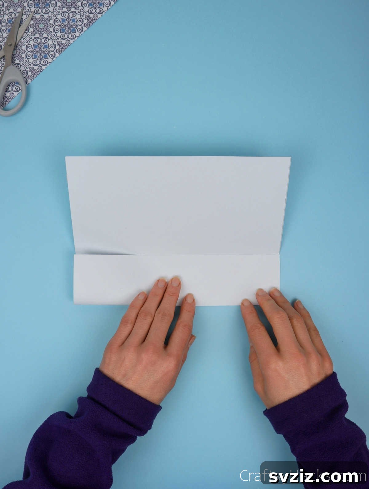 The bottom half of a paper sheet being carefully folded upwards to meet the central horizontal crease, demonstrating precise paper handling.