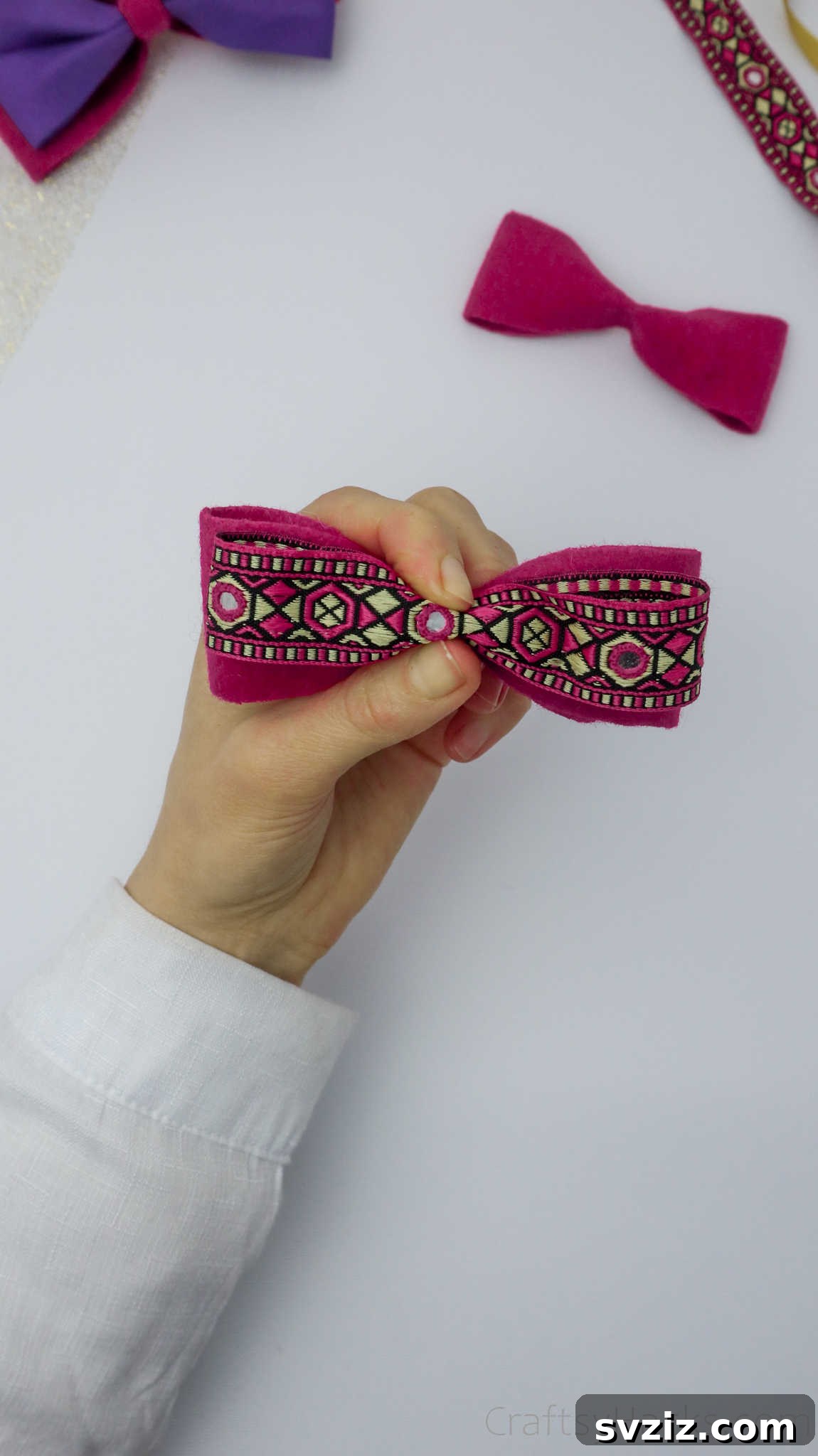 A hand adding a decorative ribbon to the center of a pink felt bow, enhancing its visual appeal