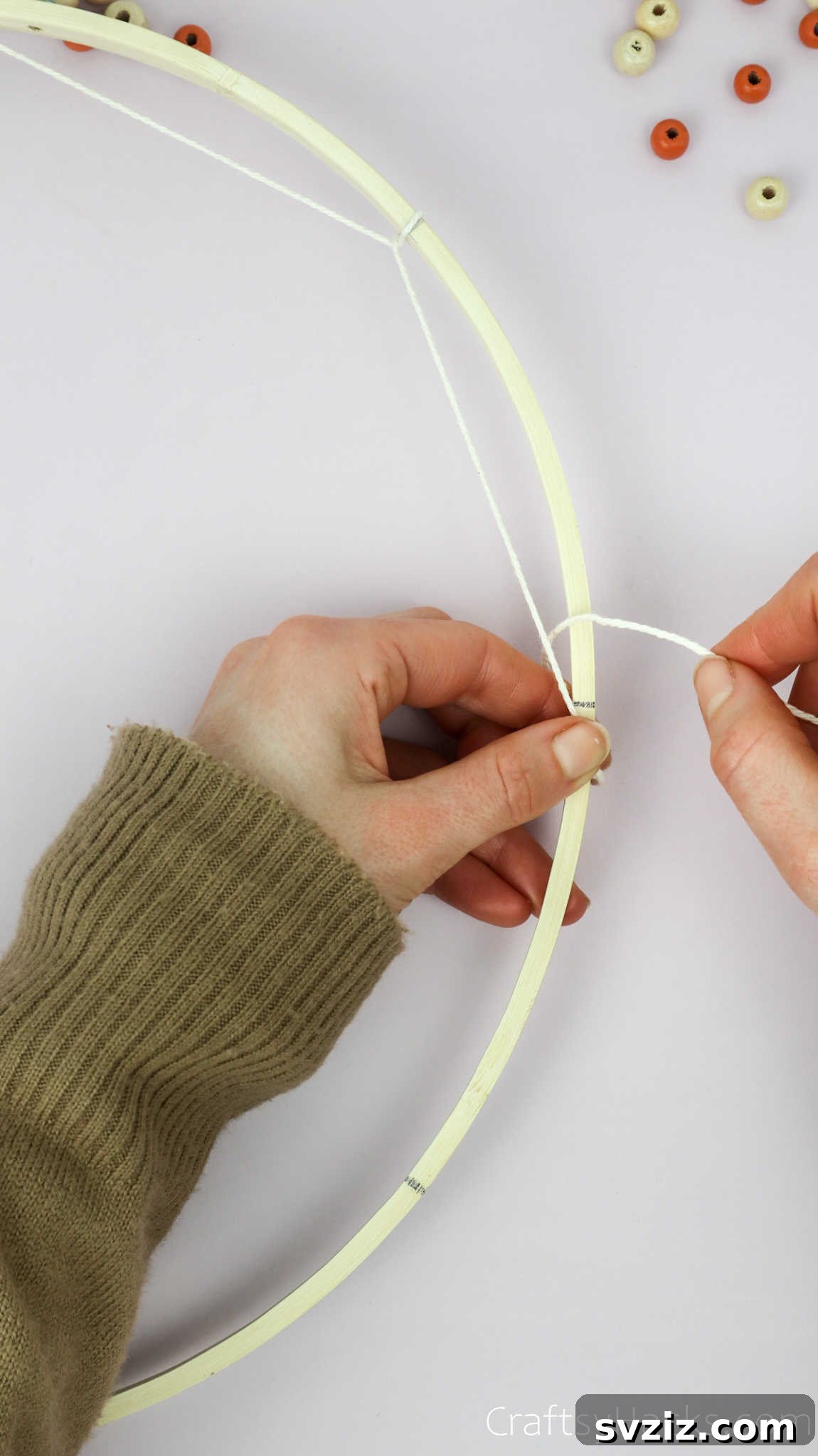 From Clutter to Charm: DIY Jewelry Storage 14 A hand continues the weaving process, looping the beige cotton string around the next marked point on the embroidery hoop, building the first layer of the dreamcatcher web.