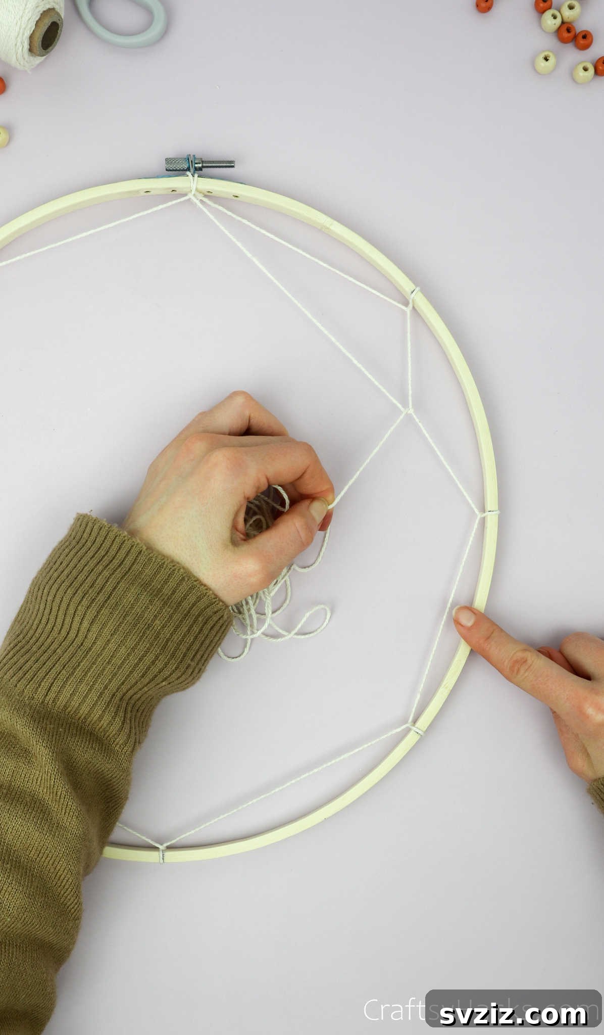 From Clutter to Charm: DIY Jewelry Storage 17 A hand starting the second round of weaving, looping the beige cotton string around the first segment of the previous string layer, moving inwards towards the center of the hoop.