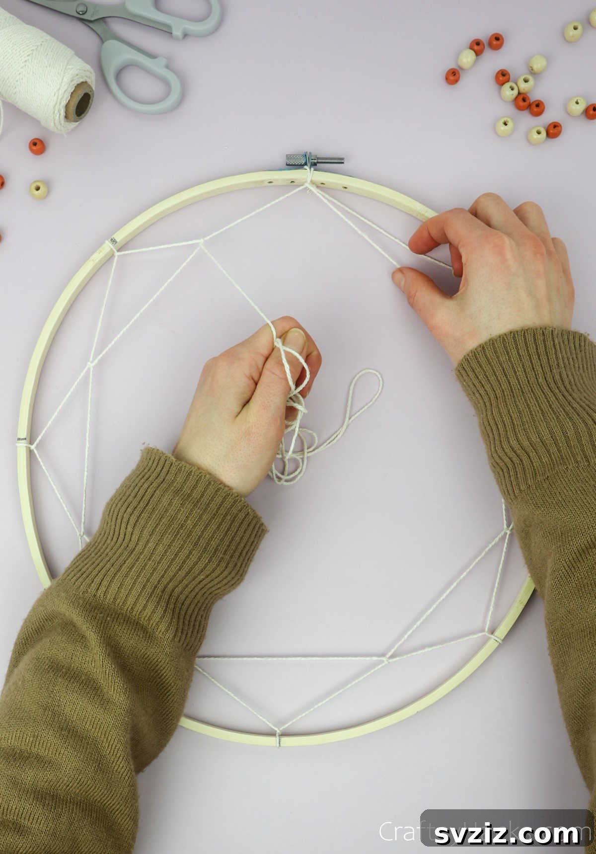 From Clutter to Charm: DIY Jewelry Storage 19 A hand continues the weaving, looping the beige cotton string around an upper segment of the previously created string layer, illustrating the progression of the dreamcatcher pattern.