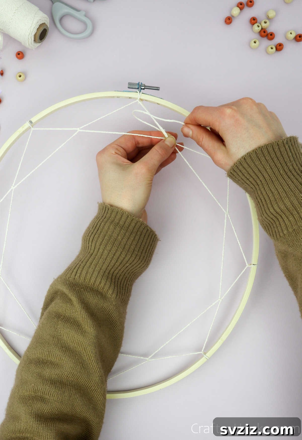 From Clutter to Charm: DIY Jewelry Storage 20 A hand attaching a new segment of beige cotton string to an existing loop within the dreamcatcher web, continuing the intricate weaving pattern.