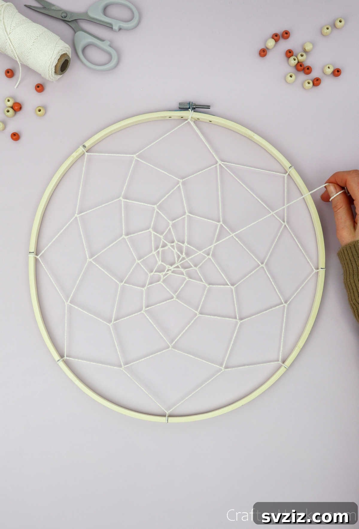 From Clutter to Charm: DIY Jewelry Storage 24 A hand tying off the beige cotton string in the middle of the dreamcatcher web, securing the final loop and completing the intricate pattern.