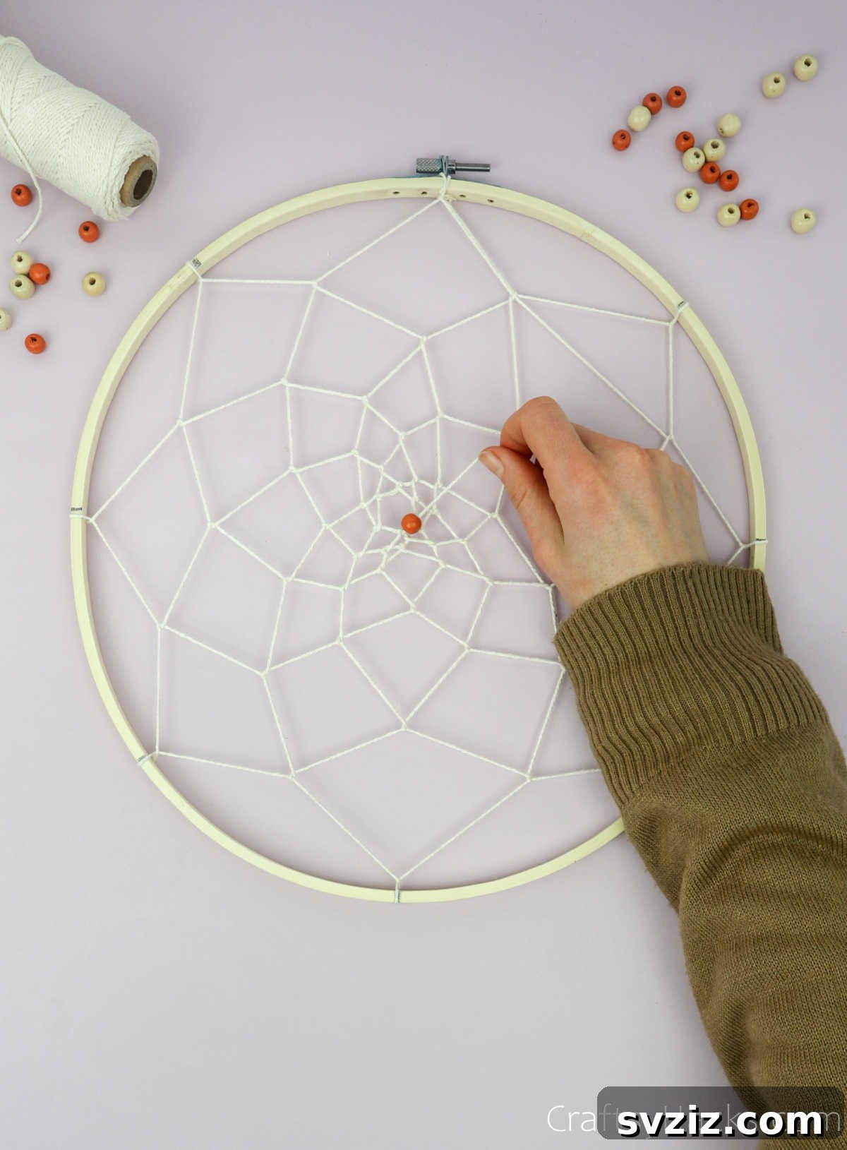 From Clutter to Charm: DIY Jewelry Storage 25 A hand carefully threading a small, round bead onto the beige cotton string at the center of the dreamcatcher web, just before tying the final knot.