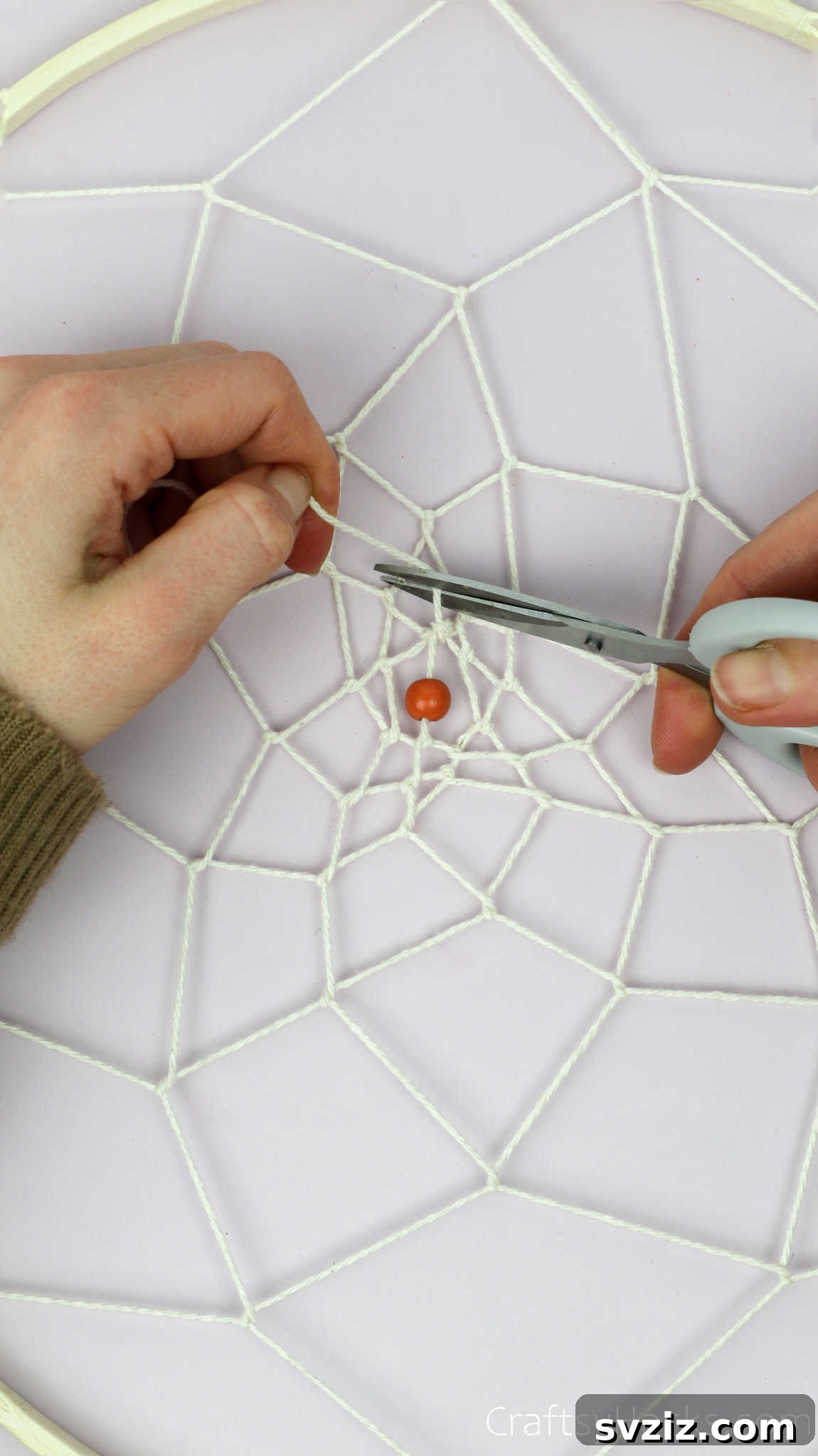 From Clutter to Charm: DIY Jewelry Storage 26 A close-up of a hand using scissors to carefully cut the end of the beige cotton string after tying the final knot in the center of the dreamcatcher web.