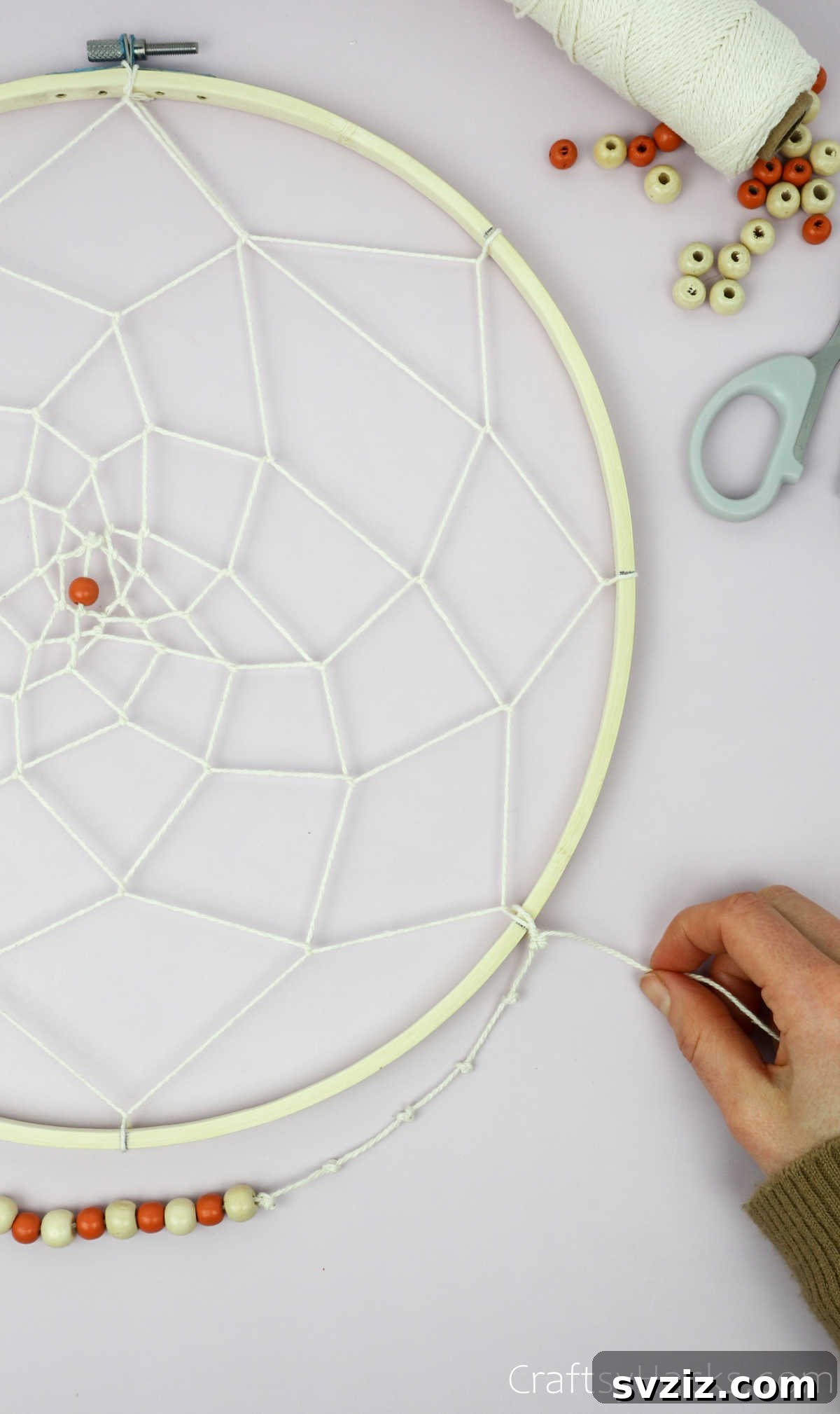 From Clutter to Charm: DIY Jewelry Storage 30 A hand tying a decorative tassel made of beige string to the bottom of the dreamcatcher's wooden hoop, adding a bohemian touch to the jewelry organizer.