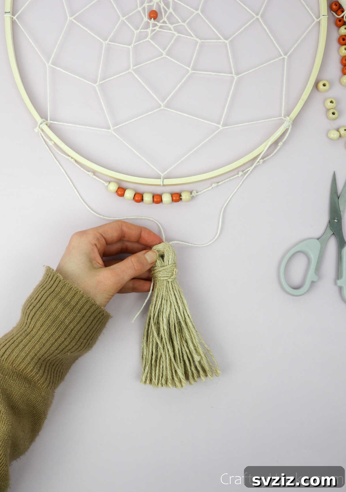 From Clutter to Charm: DIY Jewelry Storage 31 A close-up of a decorative tassel being securely tied to the bottom of the dreamcatcher's wooden hoop with a beige string, showing the final step in attaching embellishments.