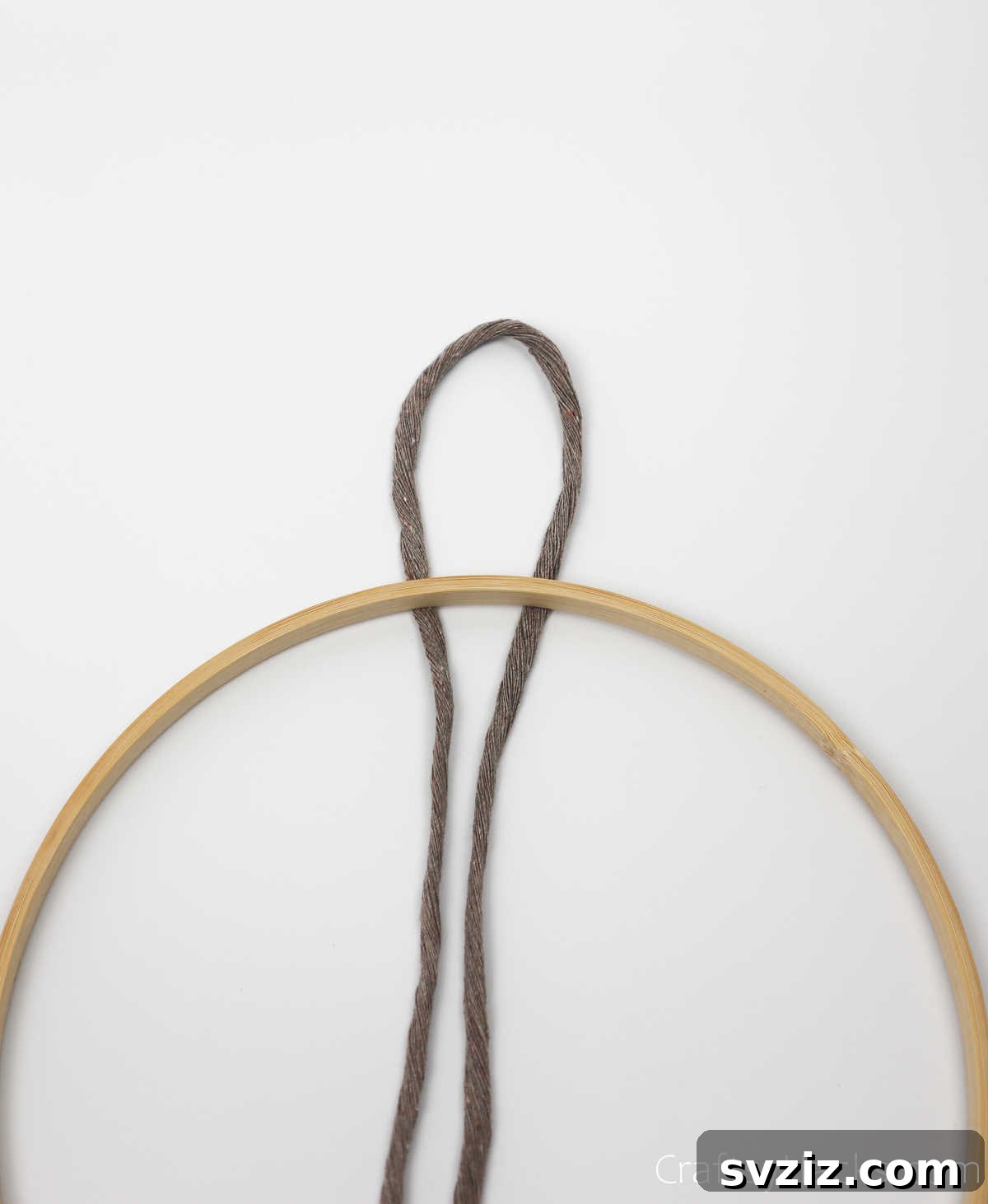 A cappuccino-colored cord being tied onto a wooden hoop, demonstrating the initial step of knotting.