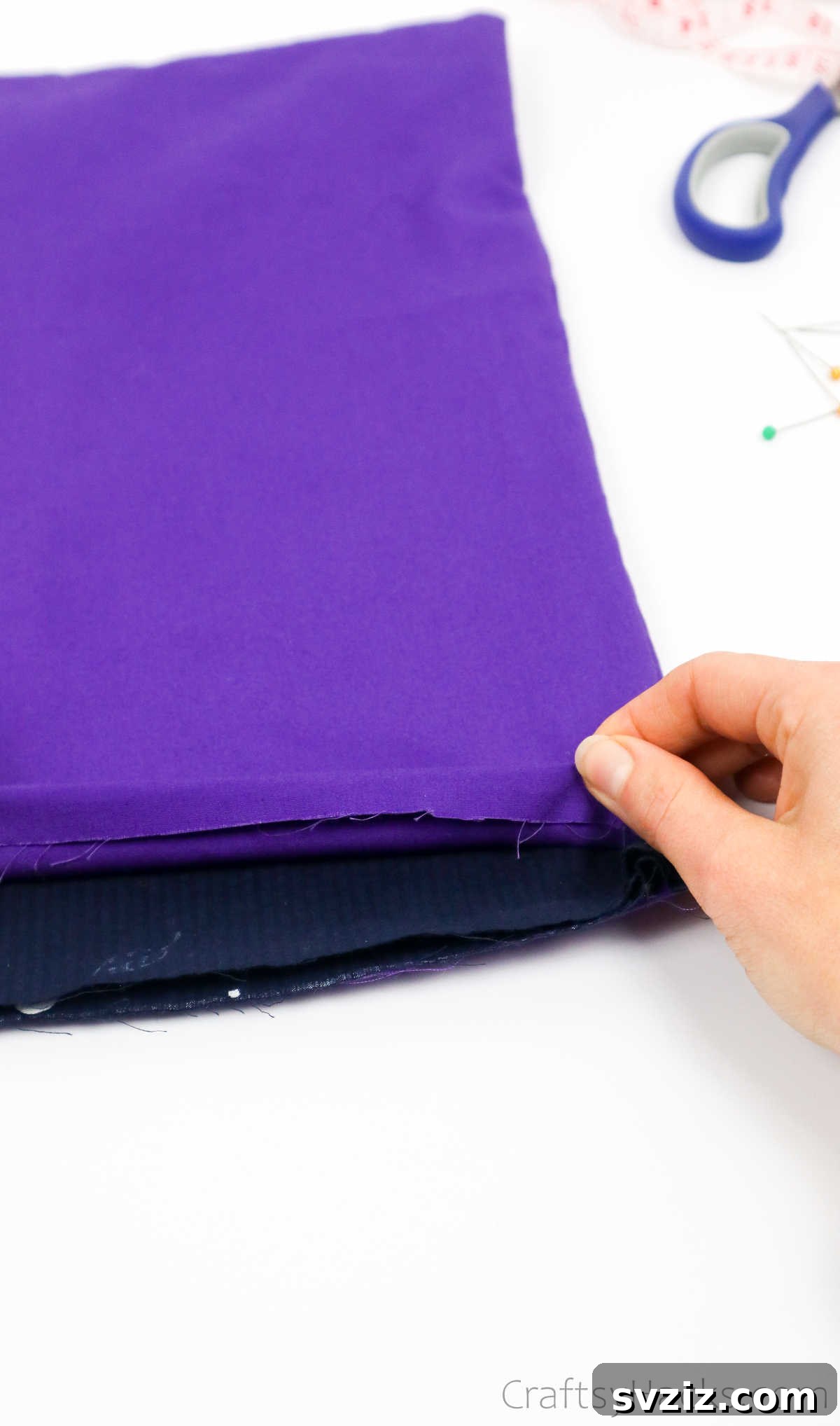 Hands folding purple fabric inwards, demonstrating the hemming process for a tote bag