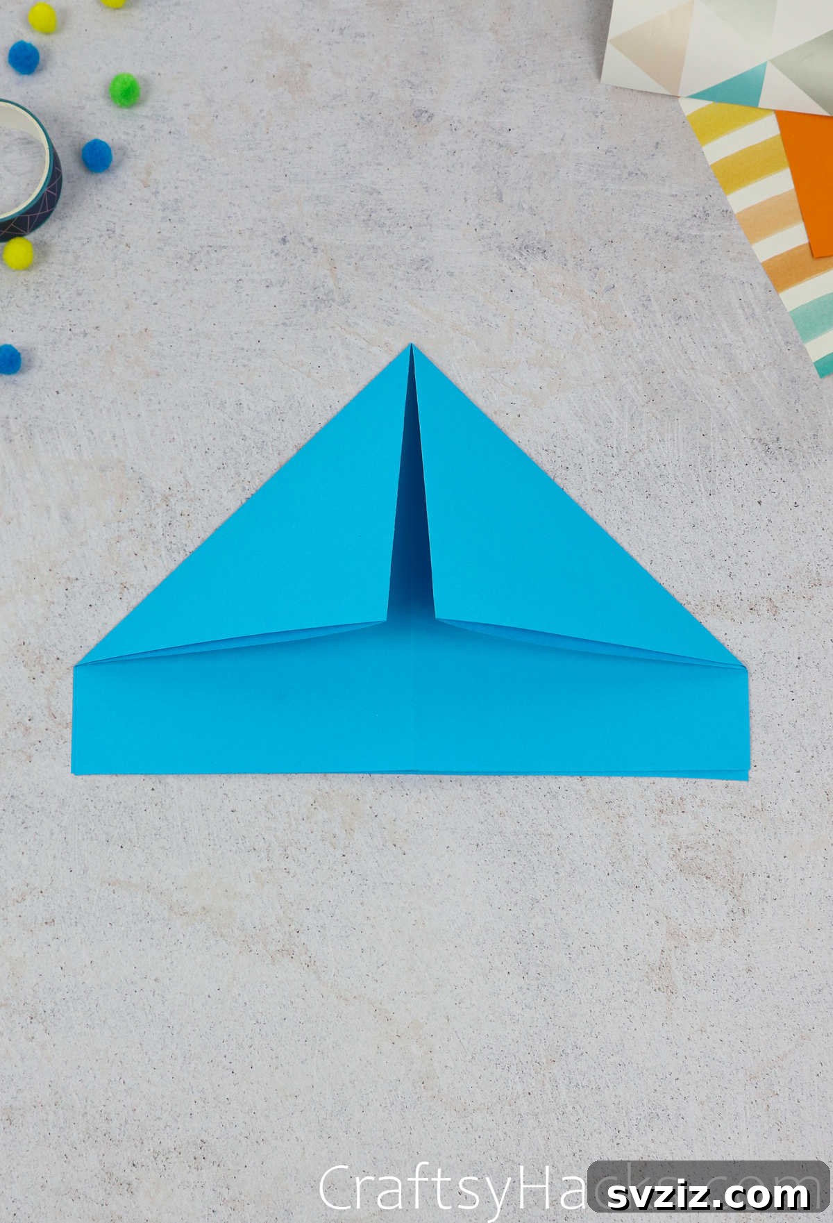 Both top corners of the blue paper are now folded down, creating a symmetrical triangular peak for the hat.