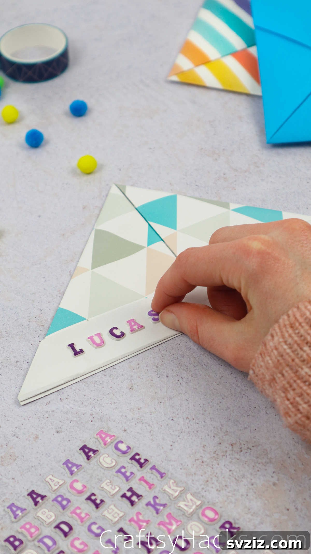 A hand adding colorful letter stickers to a finished paper hat to personalize it with a name.