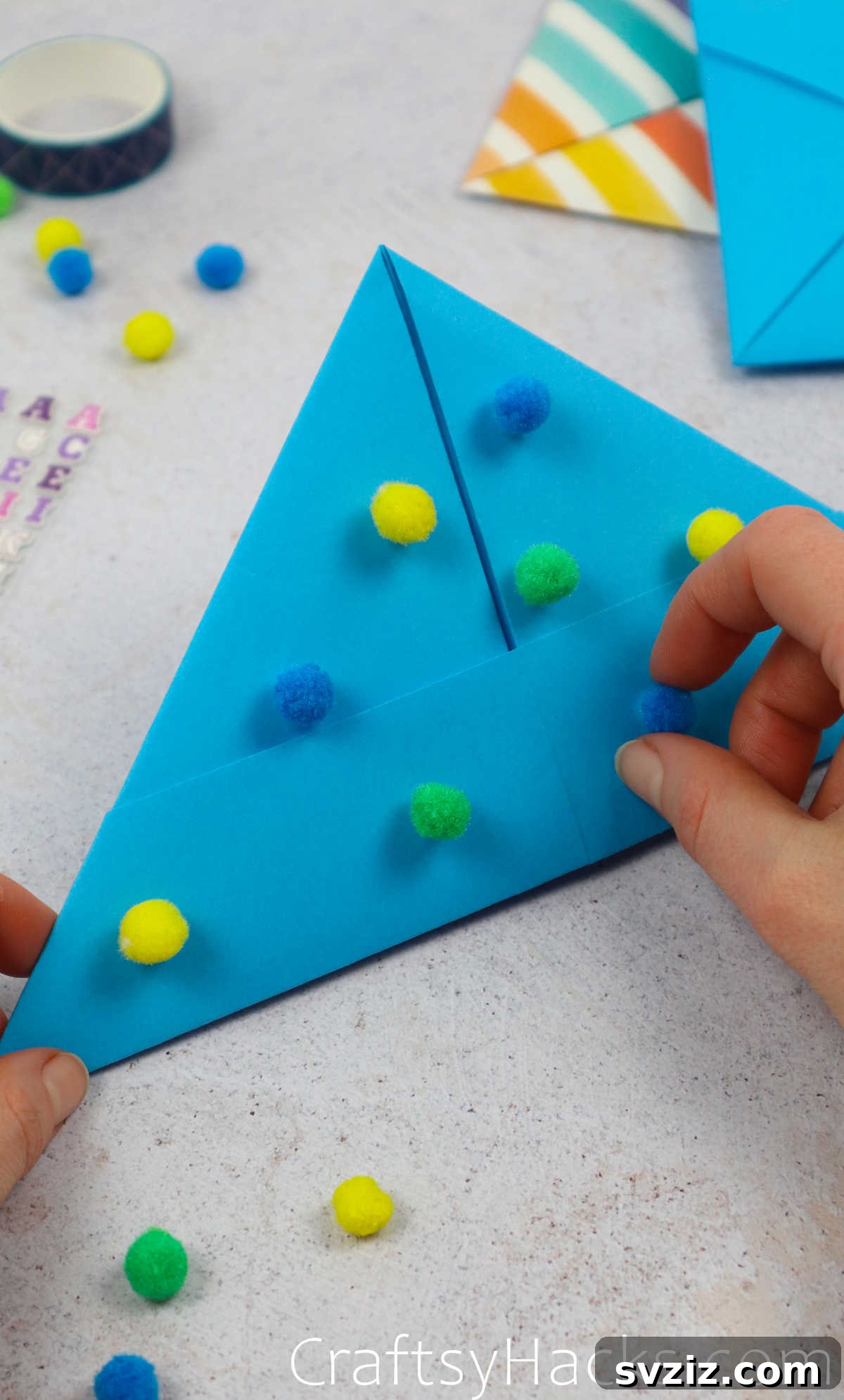 A hand carefully attaching colorful pom-poms onto a paper hat using decorative glue.
