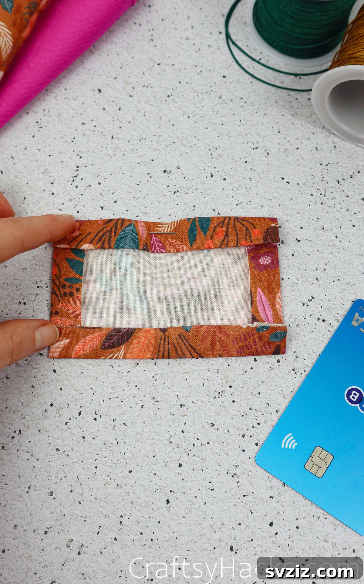 A sewing pin placed on the top edge of the folded pocket fabric, indicating the open side for card insertion.