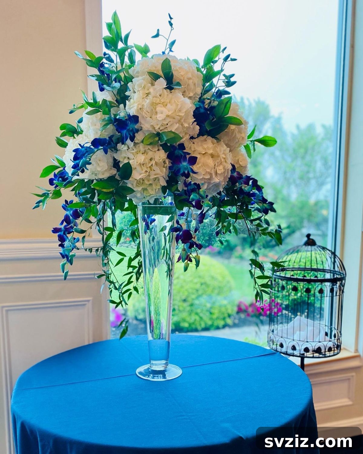 27 Elegant Round Table Wedding Centerpiece Inspirations 10 Wedding centerpiece featuring a tall V-shaped vase overflowing with white hydrangeas and blue orchids.