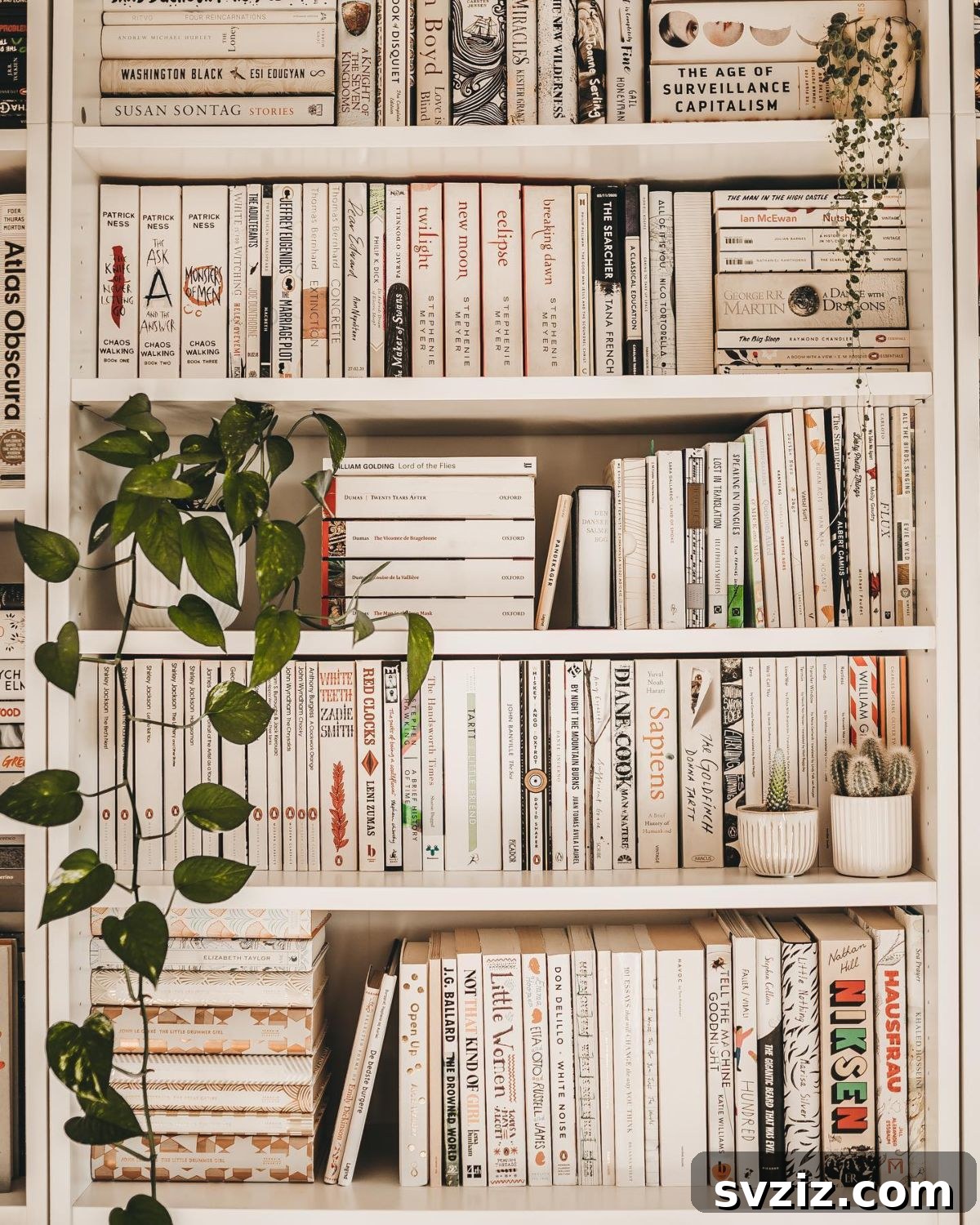All-white bookshelf with vertically and horizontally arranged books, accented by lush green plants.