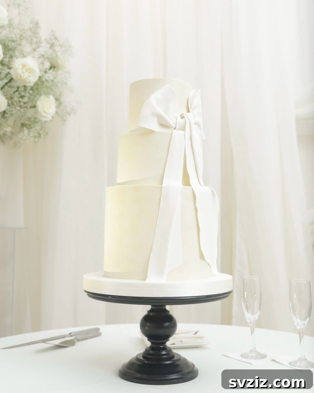 27 Elegant Wedding Cake Inspirations 11 A pure white three-tier wedding cake with a huge draped white ribbon, exuding effortless elegance and timeless beauty, presented on a contrasting black cake stand.