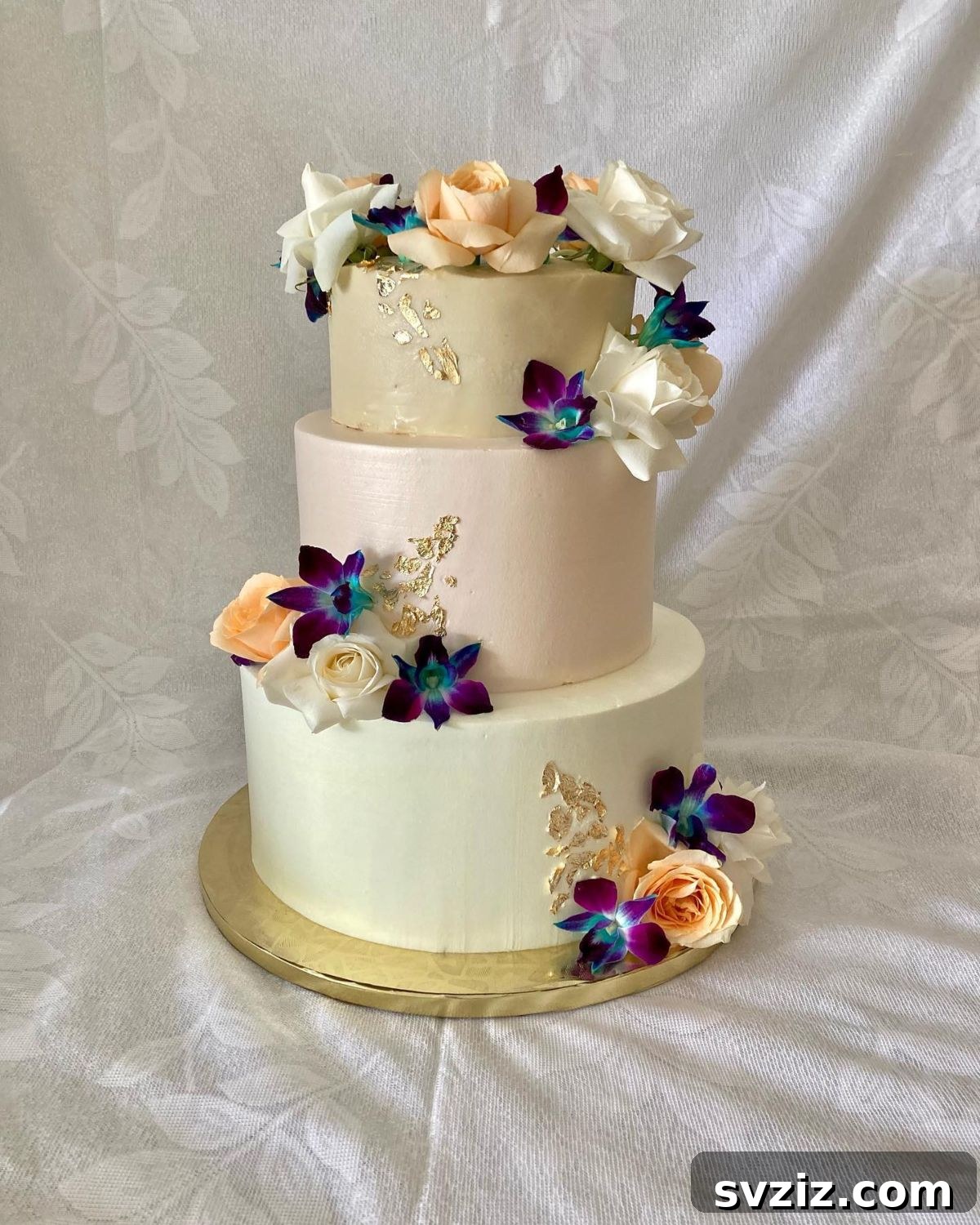 27 Elegant Wedding Cake Inspirations 14 A glorious three-tier wedding cake for the modern bride, coated in smooth buttercream and adorned with peach roses, striking purple orchids, and gold fairy dust, presented on a luxurious gold cake board.
