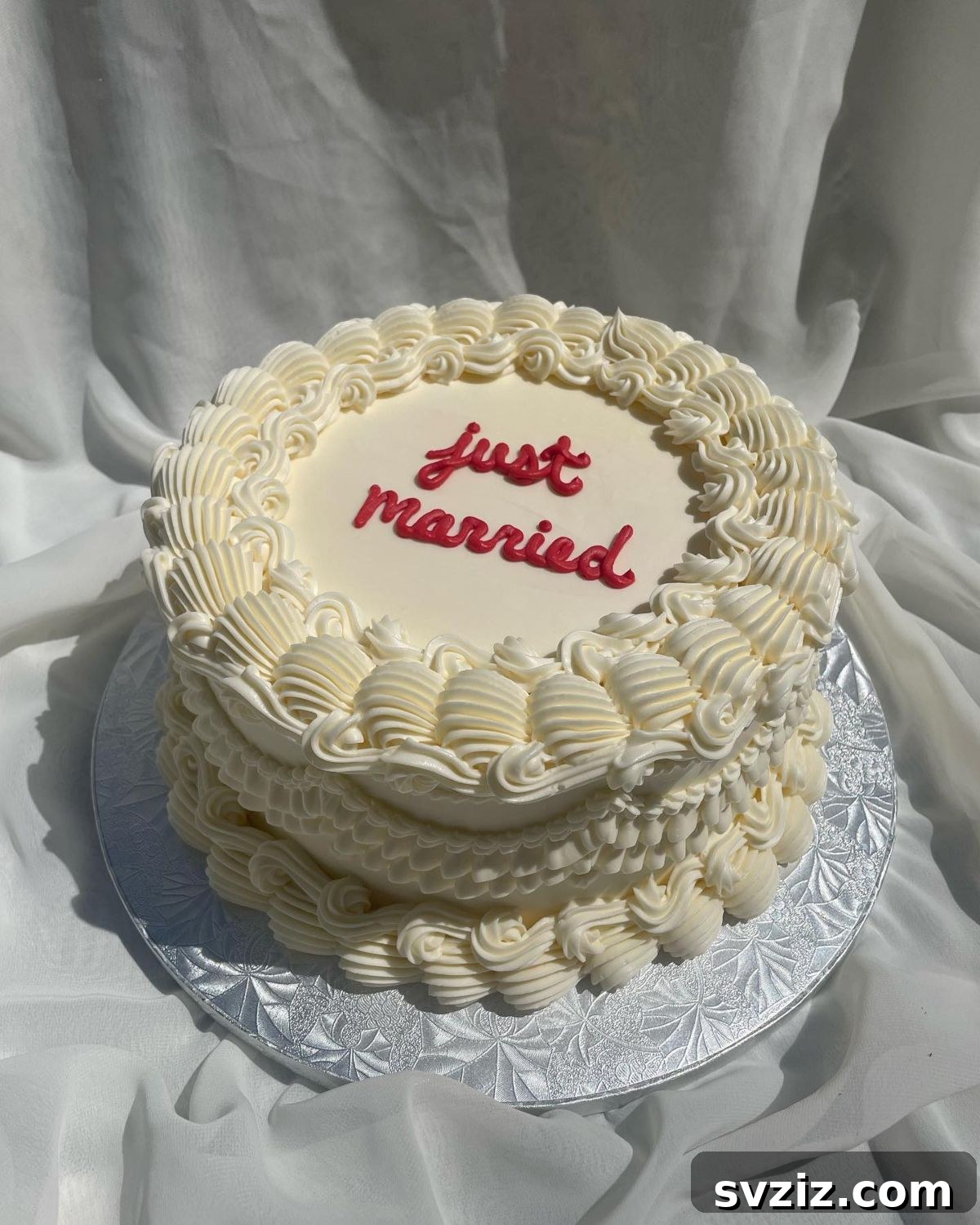 27 Elegant Wedding Cake Inspirations 4 A single-tier white wedding cake with piped shells and swirls, topped with a bold red 'Just Married' sign, showcased on a silver cake board for a modern, classy touch.