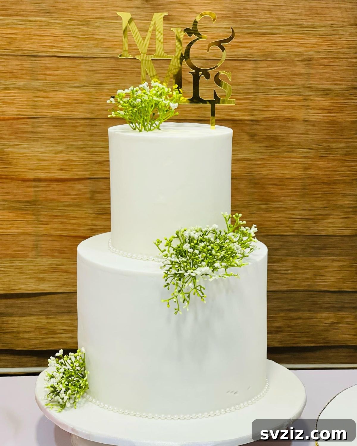 27 Elegant Wedding Cake Inspirations 8 An elegant two-tier white wedding cake adorned with tiny piped pearls, delicate sprigs of Baby’s Breath (symbolizing purity and everlasting love), and a stunning mirror gold Mr and Mrs sign as a topper.