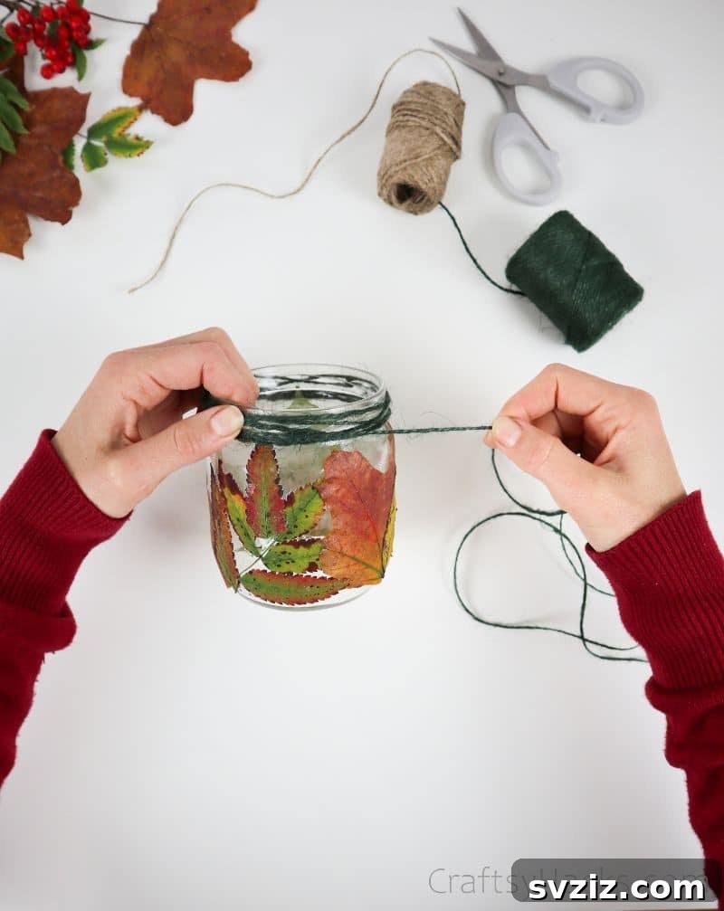 A hand wrapping green jute twine around the neck of the leaf-decorated jar, adding a decorative and concealing element.