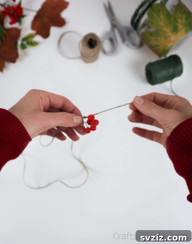 A hand tying a strand of small red decorative berries onto the jute twine wrapped around the jar's rim.