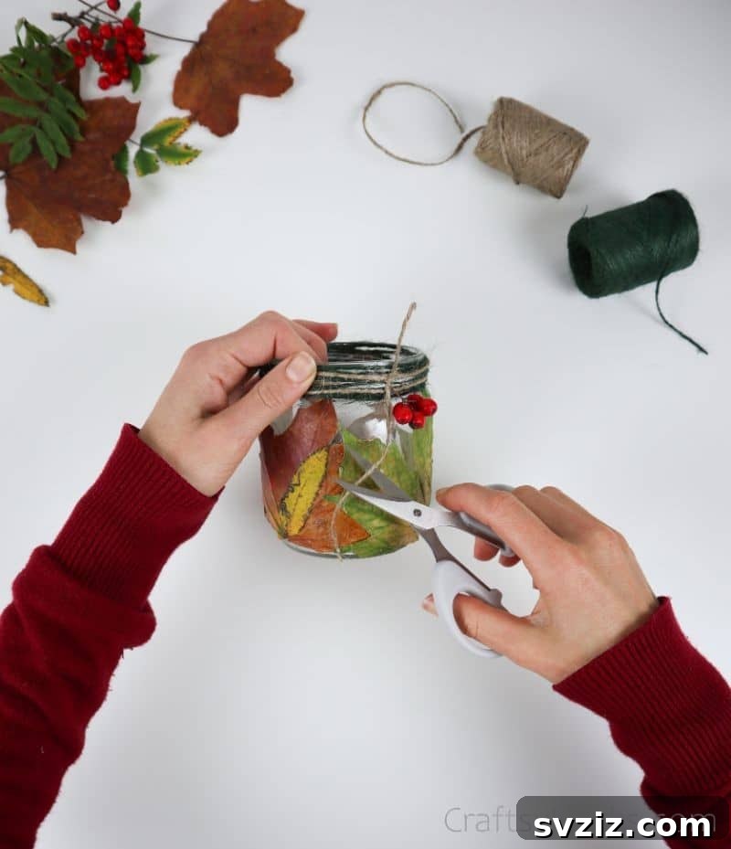 A close-up of a hand using scissors to trim the excess twine after tying decorative elements onto the jar.