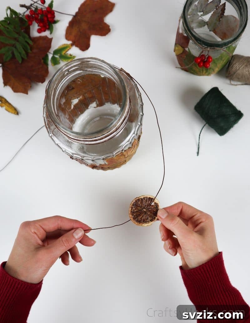 A hand threading a dried clementine slice onto a piece of twine, preparing it to be tied onto the decorated jar.